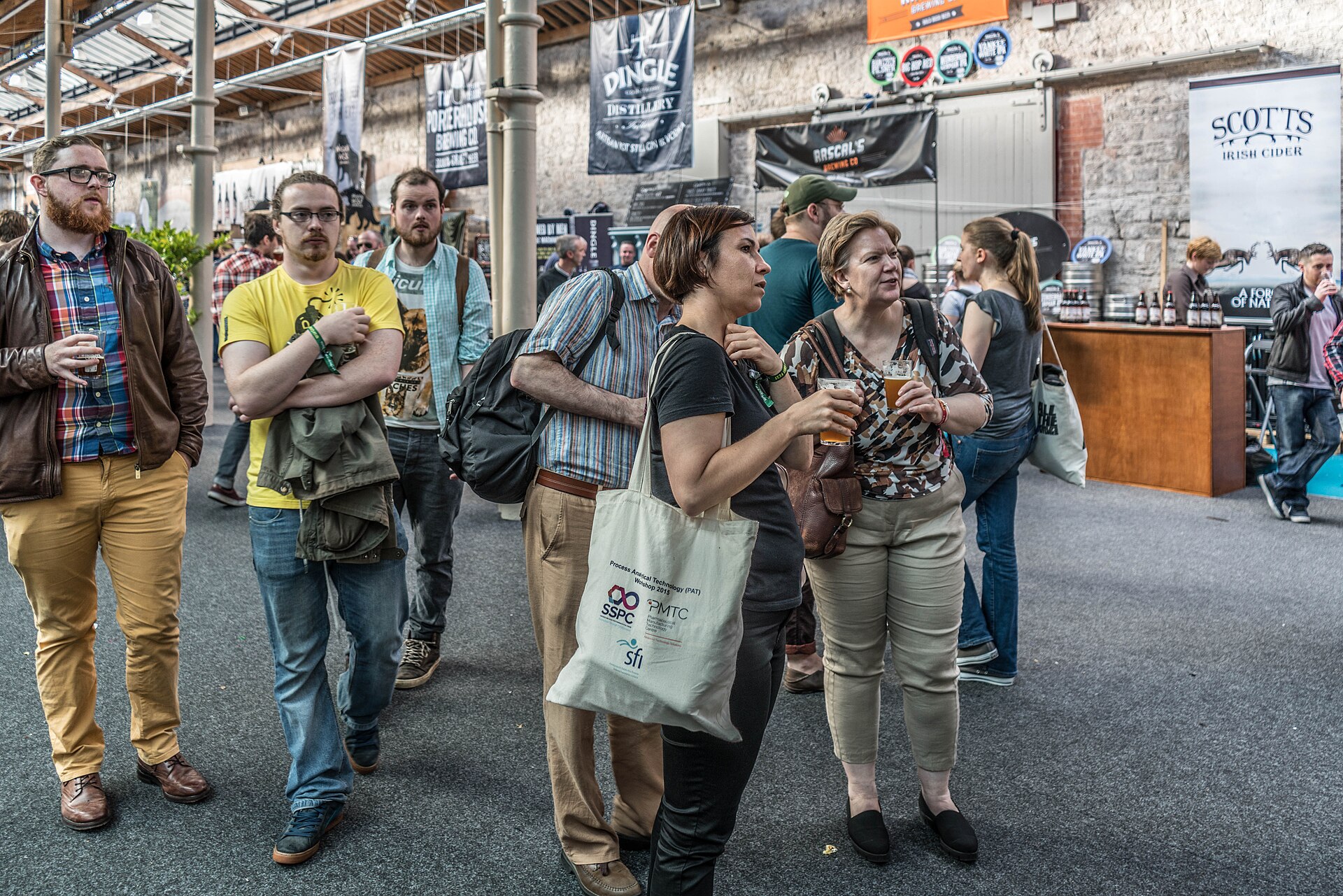 Friends walking through a bright craft beer festival hall with tasting glasses